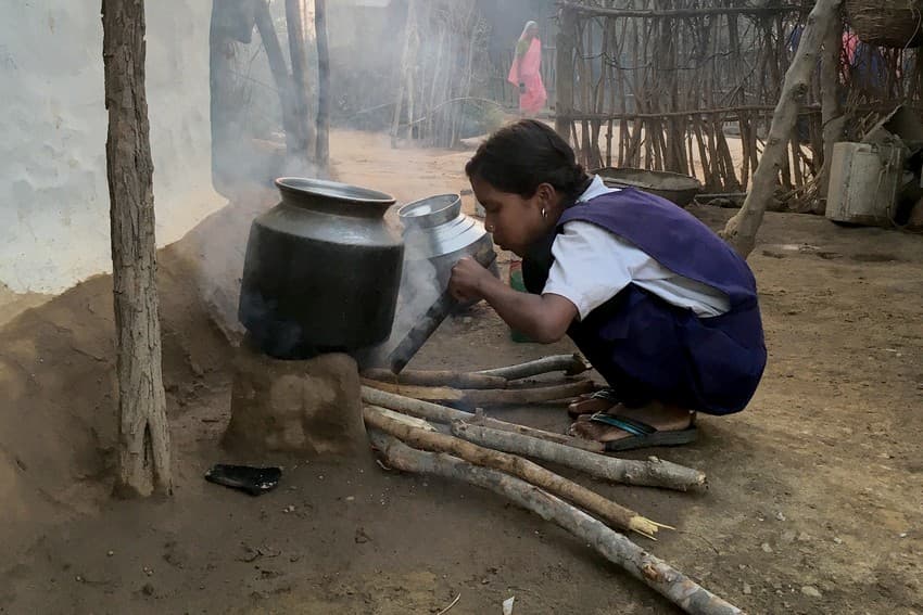 A woman uses the water heater and the fuel (crop residue) is seen in the background.